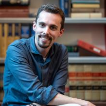 Max Chaoulideer in a blue button down shirt with rolled up sleeves in front of a bookcase full of books headshot